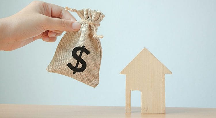 A person holding up a bag of money next to a wooden house.