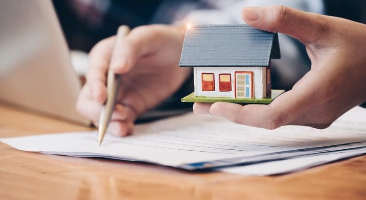 A person holding a house and pen on top of papers.