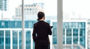 Person in office overlooking cityscape through windows.