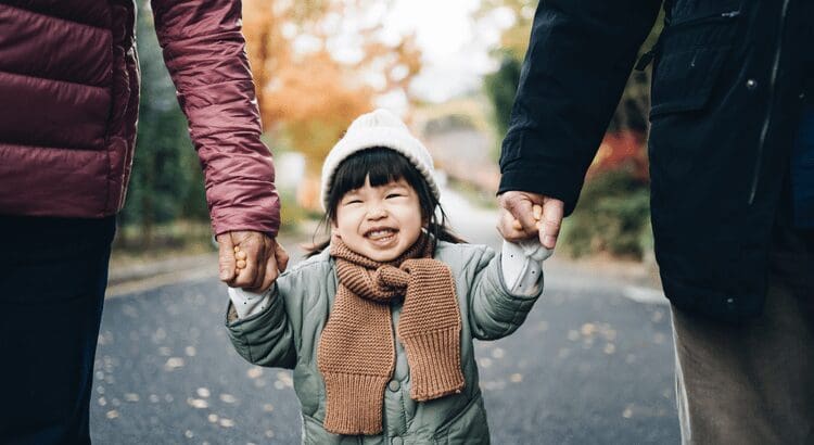 Child smiling while holding adults' hands outdoors.