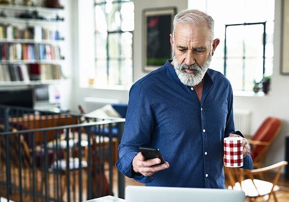 Man in his 50s in apartment, texting, with laptop on work surface, serious expression on face, planning for the day ahead, drinking coffee