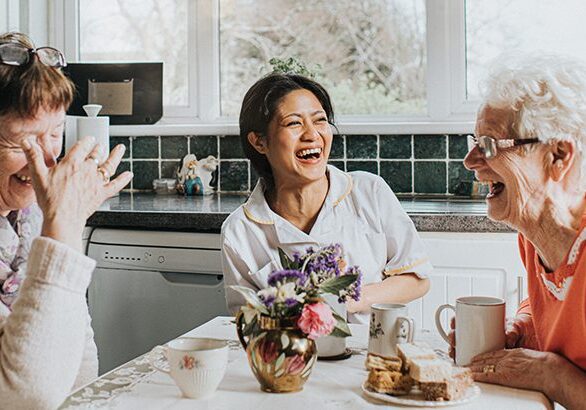 A female care assistant visits two older ladies in their home during a home visit. They sit at a kitchen table and drink tea while laughing together. They are all very comfortable and happy as they gesture and laugh loudly. The scene is upbeat and fun. Widow provides a space for copy.