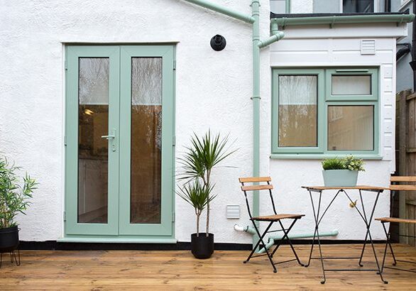 A general exterior view of a back garden patio area with wood decking, potted plants, Dragon palm tree, metal table and two chairs pale pastel sage green patio doors, window and drainpipe