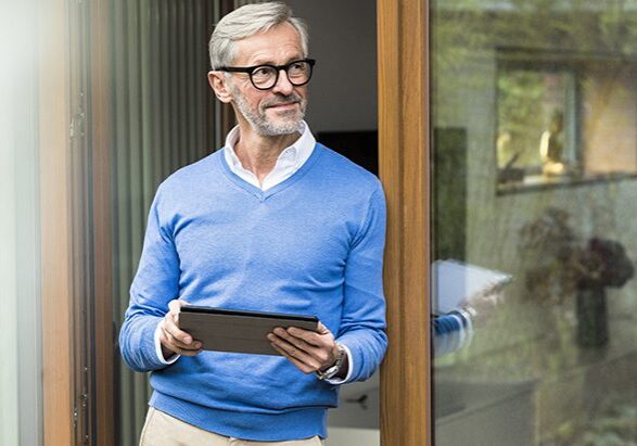 Smiling senior man with grey hair standing in front of his modern design home holding tablet, Germany