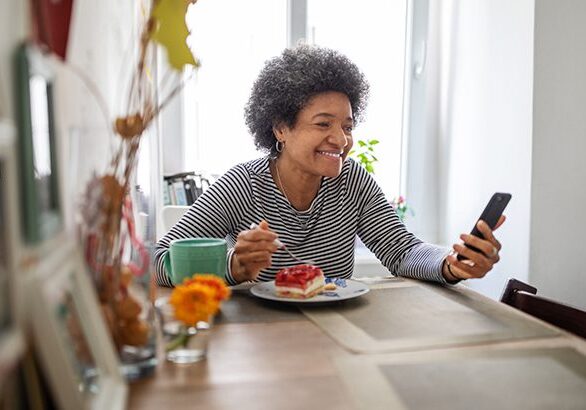 Smiling woman using smart phone and having video call during corona virus pandemic. Mature woman eating breakfast at home and having video call.