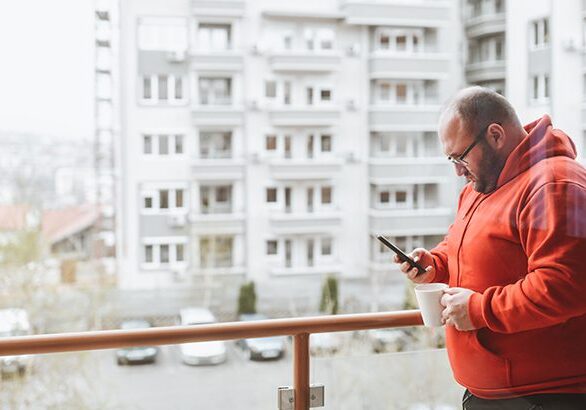 A man standing on the balcony of his apartment looking at his phone.