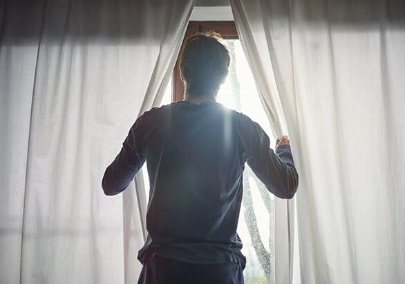 A man standing in front of a window opening curtains.