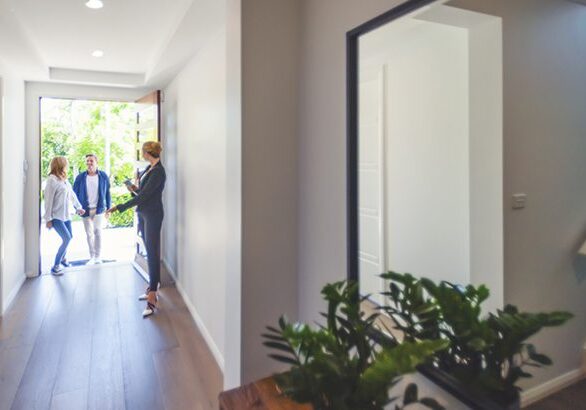 Real estate agent showing a young couple a new house. The house is contemporary. All are happy and smiling. The couple are casually dressed and the agent is in a suit. The couple are being greeted at the front door by the real estate agent. Copy space
