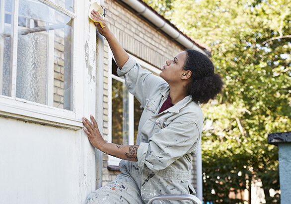 Mid adult woman scraping wall of house while standing on ladder
