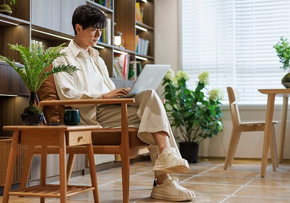 An Asian young man wearing white casual clothes. Sitting in the living room at home, working with a laptop.